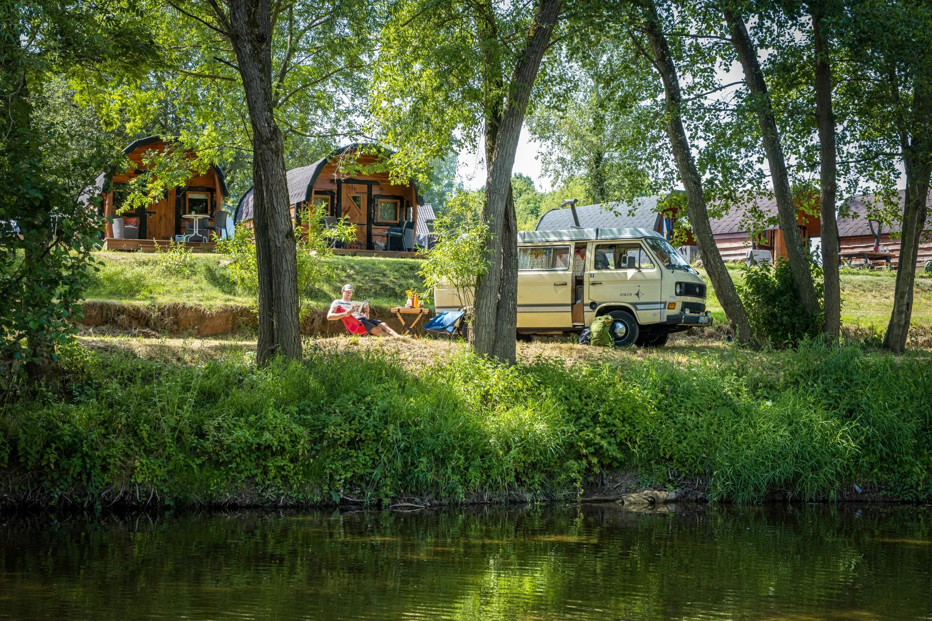 Een man ontspant in een stoel naast een vintage camperbus, met kleine houten hutjes en bomen op de achtergrond.