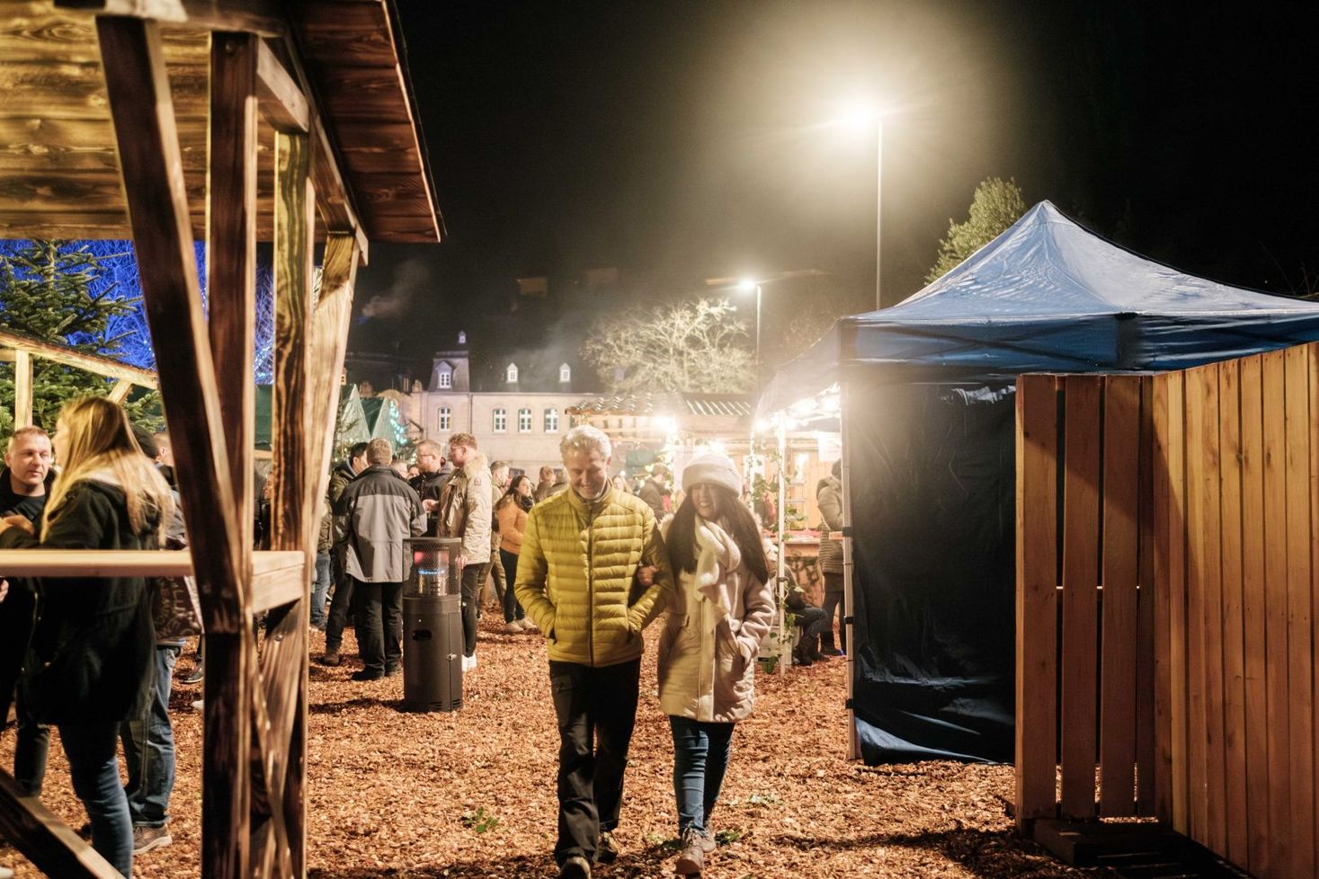 A couple walks arm in arm through the Heringer Millen Christmas market, surrounded by people, stalls, and lights.