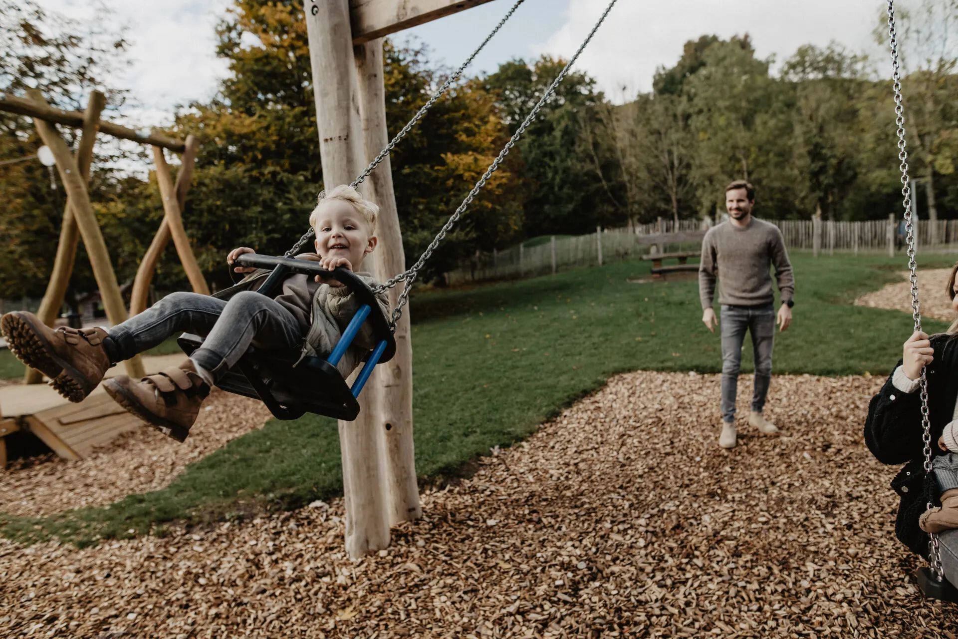 Child on a swing, mother on the right, father in the background.
