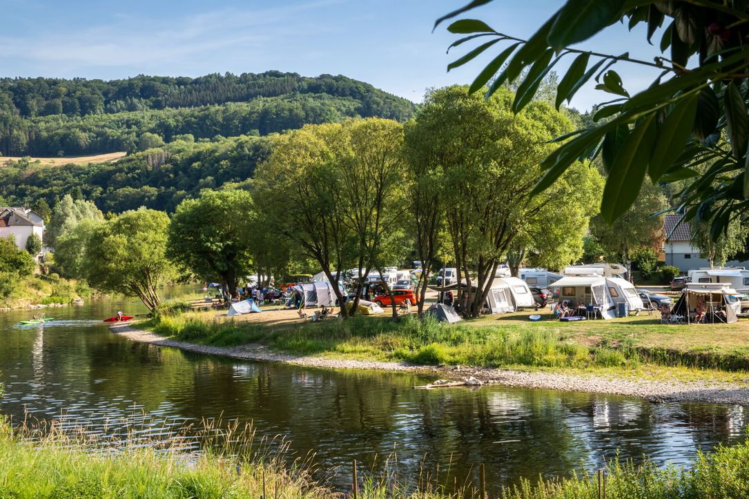 Campsite by the river, surrounded by trees and hills on a sunny day.
