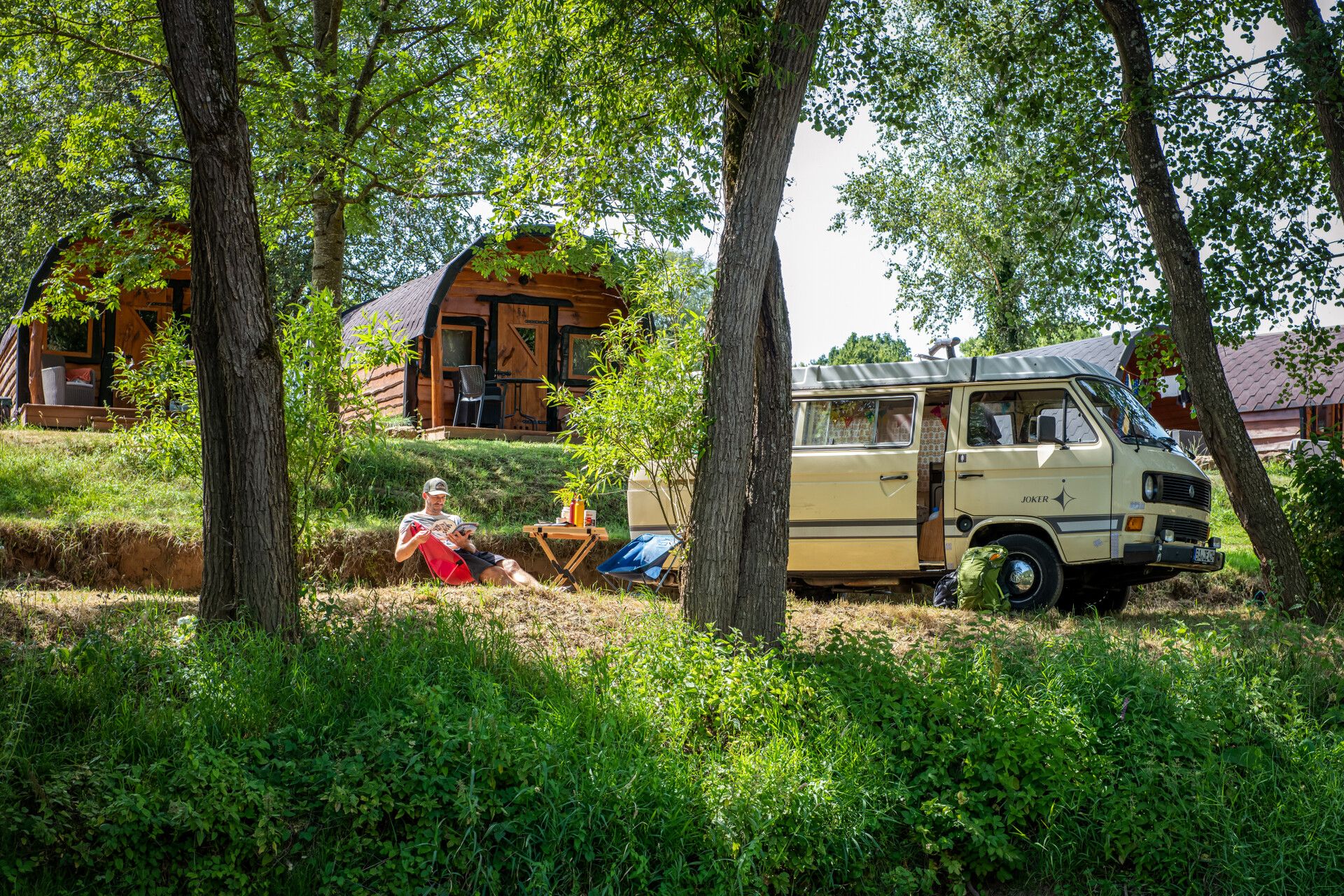 A man relaxes in a chair next to a vintage camper van, with small wooden cabins and trees in the background.