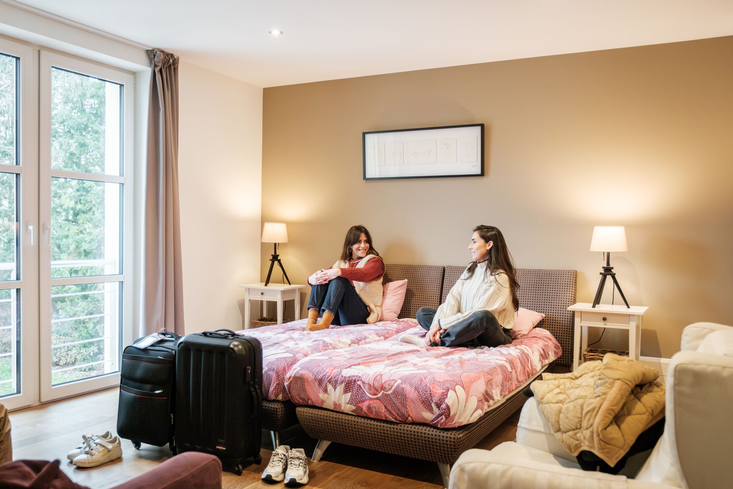 Twee vrouwen zitten met bagage op het bed in een gezellig hotelkamer.