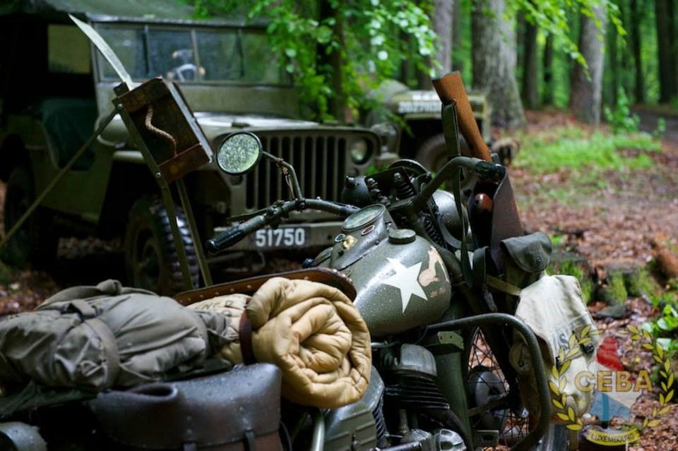 An old military motorcycle and a jeep stand in the forest, surrounded by leaves and gear.