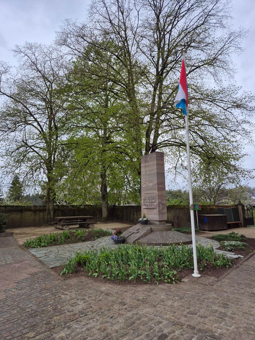 Stone war memorial with inscription and Luxembourg flag, surrounded by plants and trees.